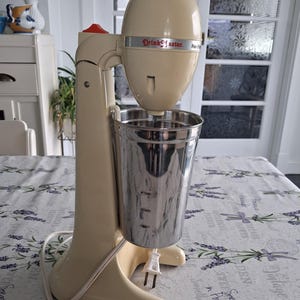 May include: A cream-colored DrinkMaster milkshake mixer featuring a tall, vertical stand with a red power switch at the top and a detachable stainless steel mixing cup. The appliance includes a white power cord with a standard two-prong plug.
