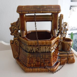 May include: A brown ceramic wishing well planter with a brick-like design. The planter has a roof, a bucket, and the words "FOR WISHING" on the front. A chain hangs from the roof to the well.