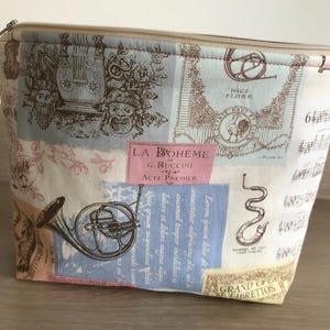 May include: A rectangular cosmetic bag with a beige zip and a fabric exterior. The fabric features a collage of musical instrument illustrations, sheet music, and text in various colours, including pink, blue, and brown.