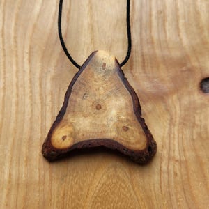 Könnte beinhalten: Ein dreieckiger Holzanhänger mit natürlichem Holzmaserungsmuster und dunkelbraunen Rindenkanten. Der Anhänger hängt an einer schwarzen Kordel. Die Holzmaserung weist einen zentralen Knoten und zwei kleinere Knoten auf. Der Anhänger ist ca. 7.6 cm breit.