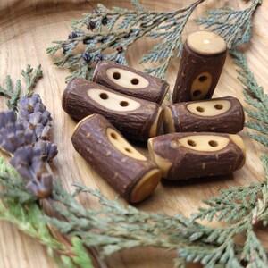 Könnte beinhalten: Eine Sammlung handgefertigter Holzknebelknöpfe. Die Knöpfe sind zylindrisch mit natürlicher Holzmaserung und zwei ovalen Löchern. Die Knöpfe sind auf einer Holzoberfläche mit Zweigen von Grün und Lavendel angeordnet.