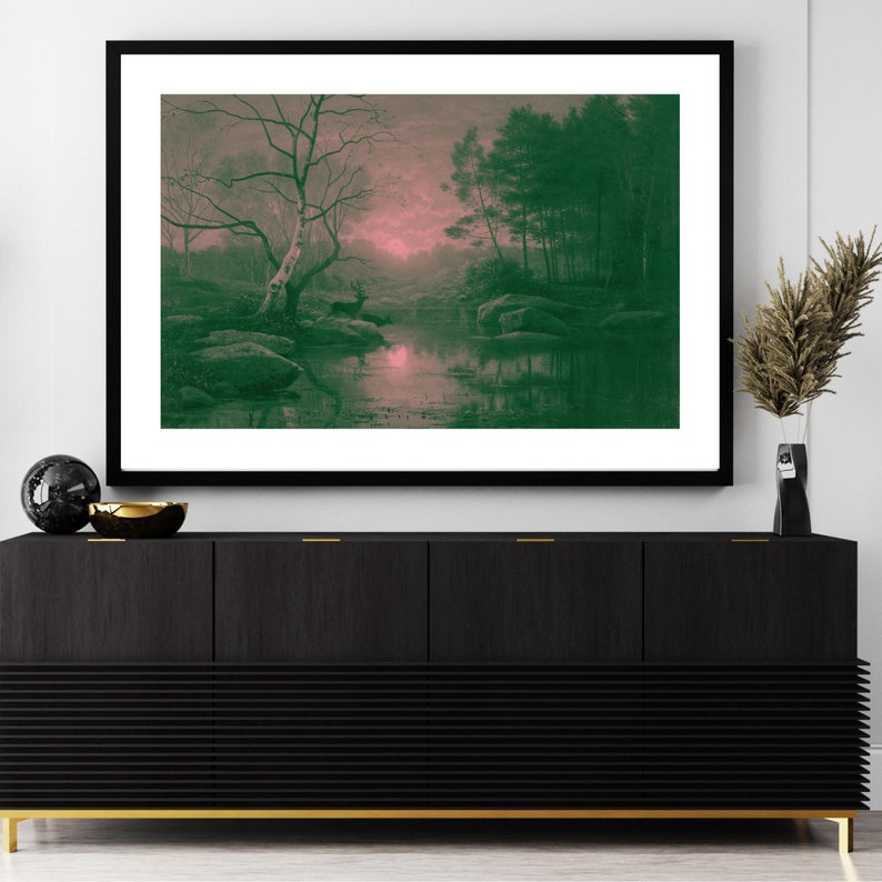 May include: Framed landscape art print featuring a serene forest scene with a deer near a body of water. The artwork has a color palette of green, pink, and gray, with a black frame. The print is displayed above a black cabinet with gold legs.
