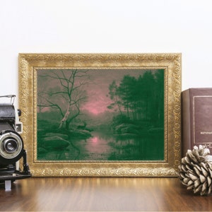 May include: A framed landscape print with a gold ornate frame. The artwork depicts a serene forest scene with a deer near a body of water. The color palette includes shades of green, pink, and brown. A vintage camera, a book, and a pinecone are on the wooden surface.