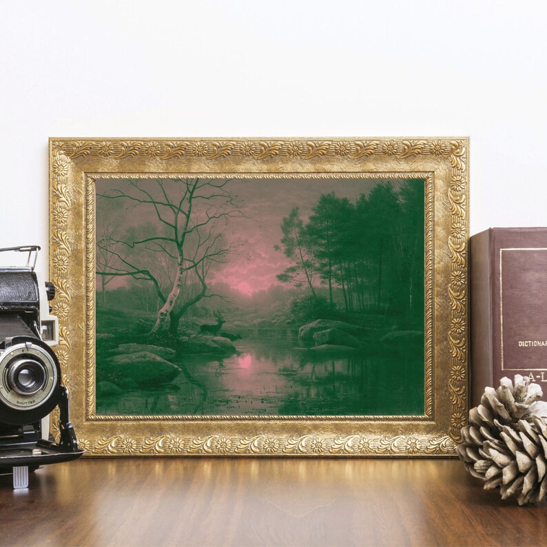 May include: A framed landscape print with a gold ornate frame. The artwork depicts a serene forest scene with a deer near a body of water. The color palette includes shades of green, pink, and brown. A vintage camera, a book, and a pinecone are on the wooden surface.