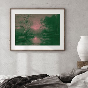 May include: A framed landscape print featuring a serene forest scene with a deer near a body of water. The artwork has a green and pink color palette, with trees, rocks, and reflections in the water. The print is in a wooden frame.
