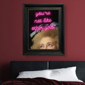 May include: A framed print of a woman's face with pink neon text that reads "you're not like other girls" above her head.