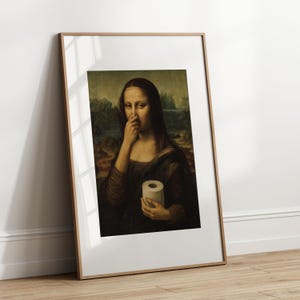 May include: Framed print of the Mona Lisa, altered to depict her pinching her nose and holding a roll of toilet paper. The artwork is in a wooden frame, leaning against a white wall and wooden floor.