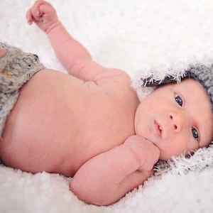 May include: A newborn baby wearing a gray and white crocheted hat with black buttons. The baby is lying on a white fluffy blanket.
