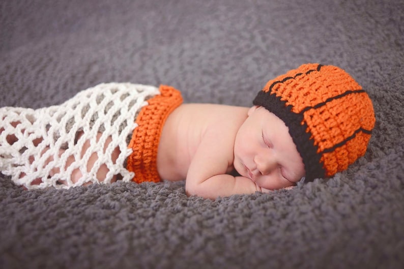 May include: A newborn baby wearing a crocheted orange and black hat and a white crocheted diaper, sleeping on a gray fuzzy blanket.