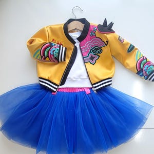 May include: A yellow satin jacket with colorful embroidered patches and a black collar, paired with a blue tulle skirt. The jacket has black and white striped cuffs and a zipper closure. A white top is visible underneath.