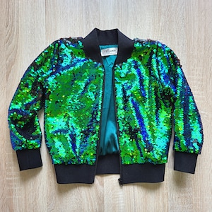 Peut inclure: Une veste bomber en sequins verts avec une fermeture éclair noire et des poignets et une ceinture côtelés noirs. La veste est doublée de turquoise et porte une petite étiquette blanche avec le mot "Byamia" dessus.