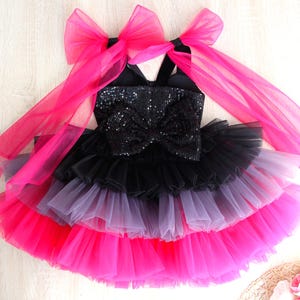 May include: A black and pink tutu dress with a sequined bodice and large bow. The dress features layers of tulle in black, grey, and bright pink. Two large pink bows are attached to the straps.