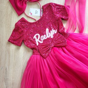 May include: A pink tulle dress with a sequined bodice and a matching pink bow. The bodice has the name "Raelyn" embroidered on it. The dress is paired with a pink headband with a large bow and a feather accent.