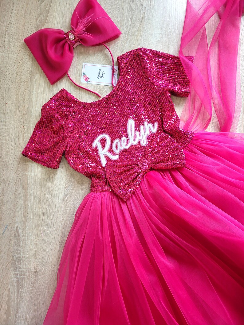 May include: A pink tulle dress with a sequined bodice and a matching pink bow. The bodice has the name "Raelyn" embroidered on it. The dress is paired with a pink headband with a large bow and a feather accent.