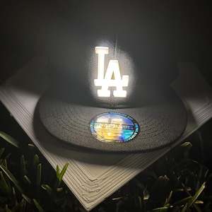 Può includere: Un cappellino snapback nero con il logo "LA" bianco. Il cappellino ha una visiera piatta e un adesivo circolare con le parole "Original Snapback Cap". Il cappellino è appoggiato su una superficie bianca.