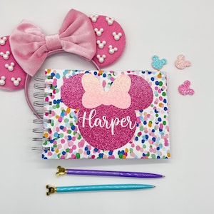 May include: A pink Minnie Mouse ear headband with white pearl accents and a matching notebook. The notebook features a pink glitter Minnie Mouse design with the name "Harper" and colorful confetti. Two pens, purple and teal, are also included.