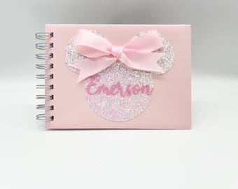 Könnte beinhalten: Ein rosa Spiralnotizbuch mit einem Design im Minnie Mouse-Stil. Das Design zeigt glitzernde Ohren, eine rosa Schleife und den Namen "Emerson" in rosa Schrift. Das Notizbuch befindet sich auf weißem Hintergrund.