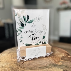 May include: A white card with green watercolor leaves and the text "do everything in love 1 COR. 16:14" printed on it. The card is on a wooden stand with a small white tag that says "Standfast Truths" and "seven hand-lettered bible verse prints on heavy card stock, with a natural oak display stand. Holly Letters."