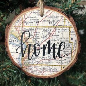 May include: A round wooden Christmas ornament with a map design and the word "home" in black script. The ornament has a natural wood edge and is suspended by a twine loop. The map features towns and roads.
