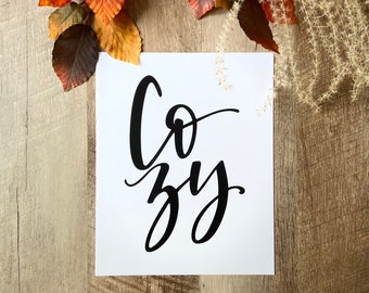 May include: A white printable sign with the word "Cozy" written in black calligraphy. The sign is surrounded by fall leaves and dried grasses.