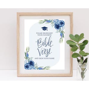 May include: A framed print with a light wood frame. The print features blue floral accents and text that reads "PLEASE HIGHLIGHT YOUR FAVORITE Bible Verse AND SIGN YOUR NAME". A graduation cap graphic is at the top.