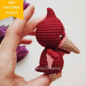 May include: A crocheted amigurumi bird toy in red and pink with a long, beige beak. The bird is being held in a hand.