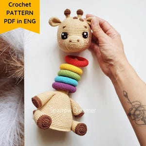 Op de afbeelding: Een gehaakt girafspeeltje met een stapel kleurrijke ringen voor de nek. De giraffe heeft een bruin lichaam, bruine vlekken en een schattig gezicht. De afbeelding bevat de tekst "Crochet PATTERN PDF in ENG".