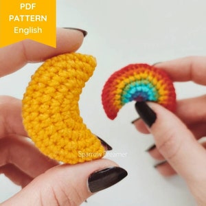 May include: Two crocheted amigurumi charms, a yellow crescent moon and a rainbow, are held in the hands of a person. The charms are small and detailed, with a textured surface. The text "Sparrow Dreamer" is visible in the image.