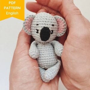 May include: A crocheted koala amigurumi toy with gray fur, pink ears, and black eyes. The toy is being held in a person's hands.