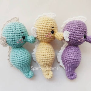 Op de afbeelding: Drie gehaakte zeepaardjes in pastelkleuren: turquoise, lichtgeel en lila. Elk heeft een witte franje, zwarte ogen en roze wangen. Zachte knuffels voor kinderen of als decoratie, met zorg gemaakt.