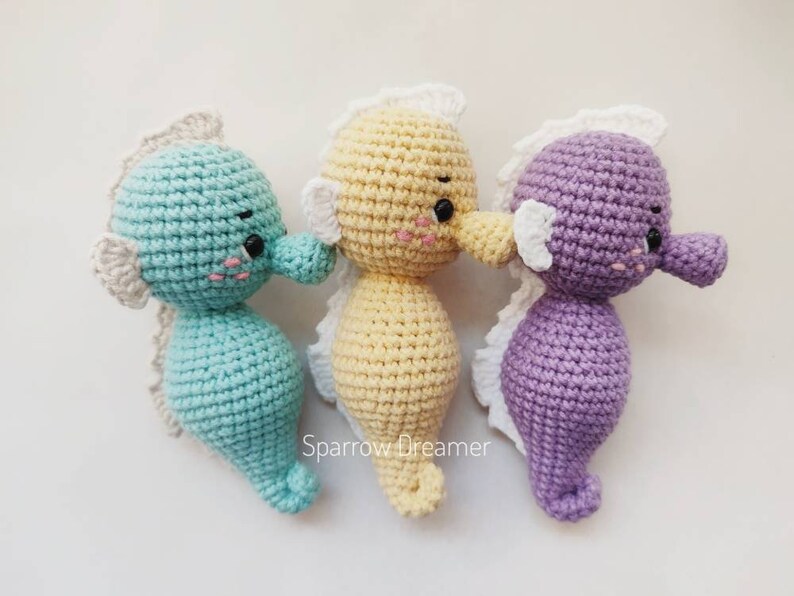 Op de afbeelding: Drie gehaakte zeepaardjes in pastelkleuren: turquoise, lichtgeel en lila. Elk heeft een witte franje, zwarte ogen en roze wangen. Zachte knuffels voor kinderen of als decoratie, met zorg gemaakt.