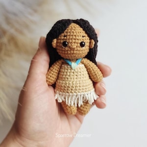 May include: A handmade crochet doll with dark hair and a tan complexion. The doll wears a light tan dress with a white fringe and a blue necklace. The doll is approximately 4 inches tall. The doll is a handmade item.