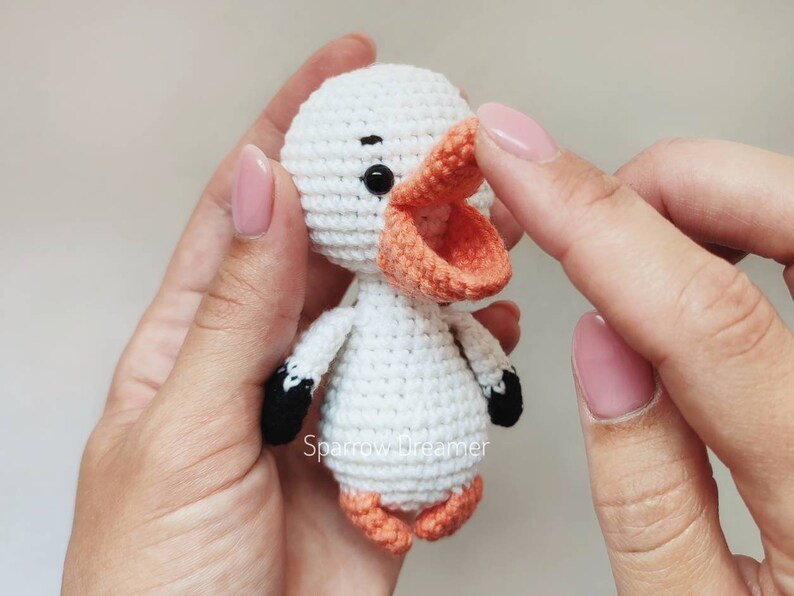 Op de afbeelding: Een handgemaakte witte gehaakte eend met een oranje snavel en poten, en zwarte accenten op de vleugels. De eend heeft een rond lichaam en hoofd, en kleine zwarte ogen. De tekst "Sparrow Dreamer" is zichtbaar.