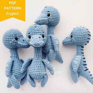 May include: Four crocheted blue dinosaur toys with black eyes and white stitching. The dinosaurs are standing on a white surface. The text "PDF PATTERN English" is visible in the top left corner of the image.