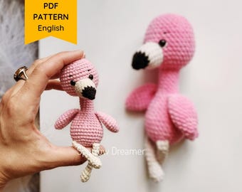May include: Two handmade pink crochet flamingo toys. One is held in a hand, the other stands on its own. The flamingos have black and white beaks and cream-colored legs. The image includes a yellow banner that says "PDF PATTERN English".