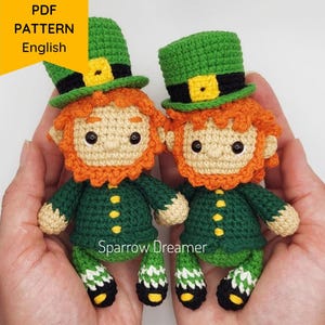 Könnte beinhalten: Zwei gehäkelte Leprechaun-Amigurumi-Puppen mit grünen Hüten, orangem Bart und grün-weiß gestreiften Hosen. Die Puppen halten sich an den Händen und werden von den Händen einer Person gehalten.