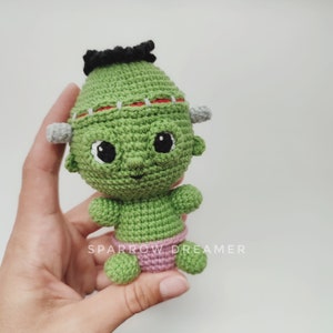 May include: Green crocheted Frankenstein monster toy with black hair, big eyes, and a pink diaper. The toy is being held in a hand.