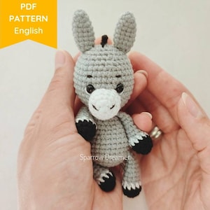 May include: A gray crocheted donkey amigurumi toy with black hooves and a white face. The donkey is being held in a person's hands.