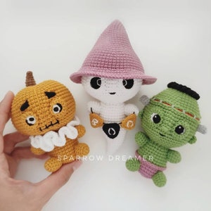 May include: Three crocheted Halloween characters: a pumpkin, a ghost wearing a pink hat, and a green monster. The pumpkin is orange with a white ruffle collar and black eyes. The ghost is white with a pink pointed hat and a black eye. The monster is green with a black headband and black eyes.