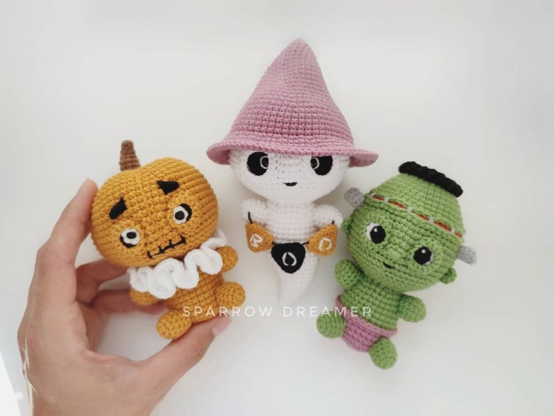 May include: Three crocheted Halloween characters: a pumpkin, a ghost wearing a pink hat, and a green monster. The pumpkin is orange with a white ruffle collar and black eyes. The ghost is white with a pink pointed hat and a black eye. The monster is green with a black headband and black eyes.