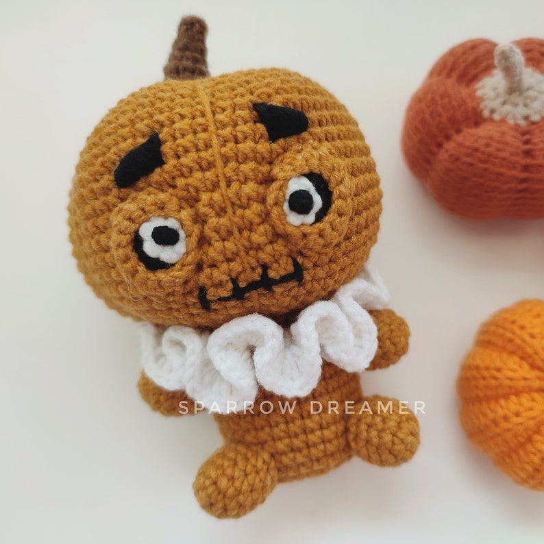 May include: A crocheted pumpkin with a white ruffle collar and a cute face. The pumpkin is orange and has black eyes and a mouth. The text "SPARROW DREAMER" is visible on the pumpkin.