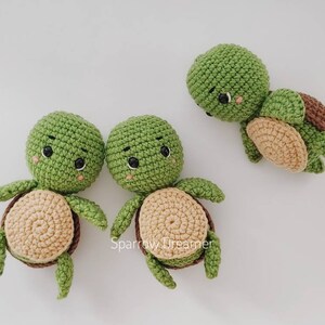 Op de afbeelding: Handgemaakte gehaakte schildpadden in groen, bruin en beige. De schildpadden hebben ronde groene hoofden, kleine zwarte ogen en lachende gezichten. De schilden zijn een mix van bruin en beige. Twee schildpadden staan rechtop en &eacute;&eacute;n ligt op zijn zij.