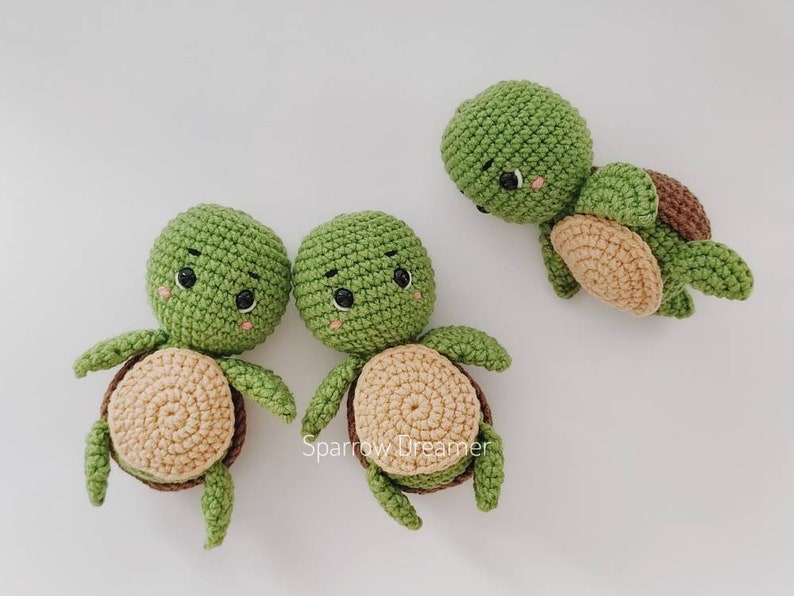 Op de afbeelding: Handgemaakte gehaakte schildpadden in groen, bruin en beige. De schildpadden hebben ronde groene hoofden, kleine zwarte ogen en lachende gezichten. De schilden zijn een mix van bruin en beige. Twee schildpadden staan rechtop en &eacute;&eacute;n ligt op zijn zij.
