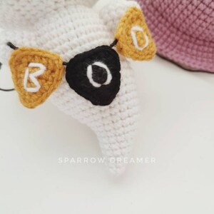 May include: White crocheted ghost with a black and white eye and a yellow crocheted banner that says "BOO" hanging around its neck.
