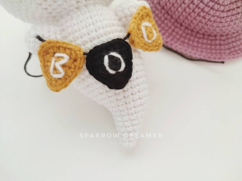 May include: White crocheted ghost with a black and white eye and a yellow crocheted banner that says "BOO" hanging around its neck.