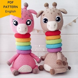 Pode incluir: Dois brinquedos de girafa de crochê com anéis de arco-íris ao redor de seus pescoços. A girafa à esquerda é rosa e a girafa à direita é marrom. As girafas estão sentadas em uma superfície de madeira branca. PDF PATTERN English.