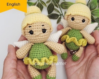 May include: Two crocheted fairy dolls with green dresses, yellow hair, and white wings. The dolls are holding hands and are being held by a person's hands.
