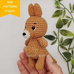 Puede incluir: Un juguete de amigurumi de canguro tejido a crochet en marrón con ojos negros y nariz blanca. El juguete está siendo sostenido en una mano. La imagen incluye el texto "PDF PATTERN English".
