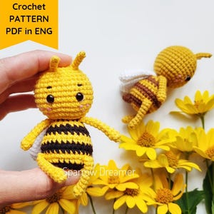 Może przedstawiać: Ręcznie robiona żółto-czarna szydełkowa pszczoła z białymi skrzydłami, trzymana w dłoni, z drugą pszczołą i żółtymi kwiatami w tle. Tekst "Crochet PATTERN PDF in ENG" znajduje się w lewym górnym rogu.
