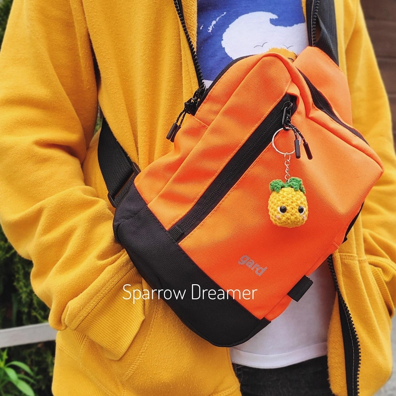 Pode incluir: Uma bolsa transversal laranja com detalhes em preto e um pequeno chaveiro de abacaxi amarelo. A bolsa tem a palavra "gard" impressa, e o texto "Sparrow Dreamer" tamb&eacute;m &eacute; vis&iacute;vel.